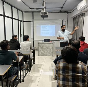 A digital marketing instructor delivering an SEO lecture to a group of students in a classroom, with a online-offline training presentation slide displayed on a projector screen.