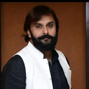 Faculty member in formal attire, wearing a black shirt with a white vest, posing against a brown background.